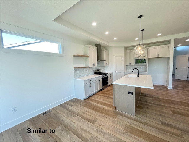 kitchen with wall chimney exhaust hood, light hardwood / wood-style flooring, stainless steel range, a tray ceiling, and a center island with sink