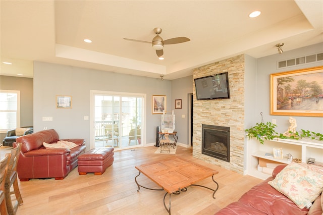 living room with a stone fireplace, a raised ceiling, light hardwood / wood-style floors, and ceiling fan