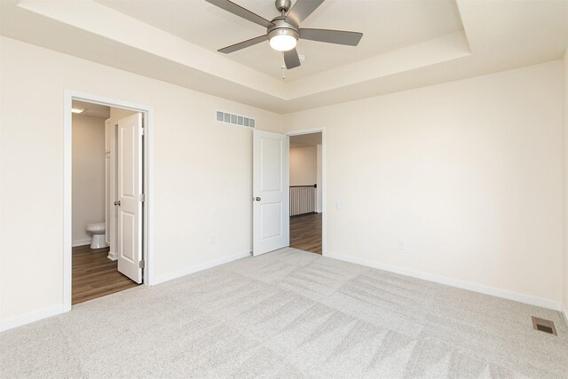 unfurnished bedroom with a raised ceiling, ceiling fan, carpet flooring, and ensuite bathroom