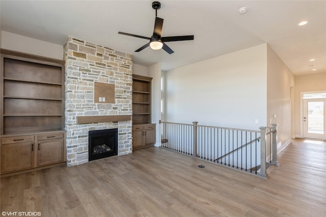 unfurnished living room with built in shelves, ceiling fan, a stone fireplace, and light hardwood / wood-style flooring