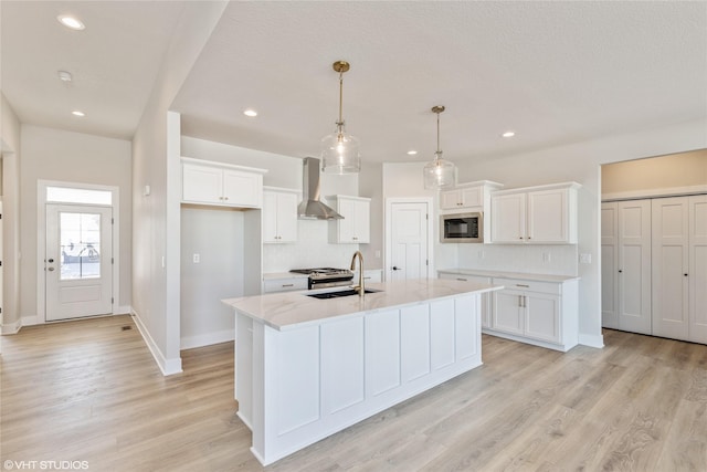 kitchen with decorative light fixtures, wall chimney exhaust hood, white cabinets, and an island with sink