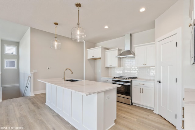 kitchen with white cabinets, wall chimney exhaust hood, an island with sink, hanging light fixtures, and stainless steel gas range oven