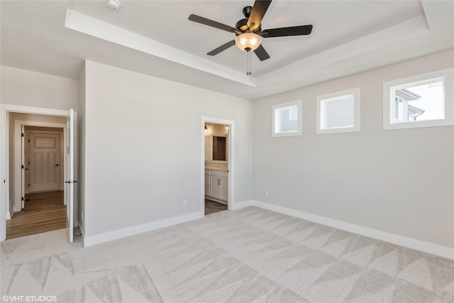 unfurnished bedroom featuring light carpet, ceiling fan, ensuite bathroom, and a tray ceiling