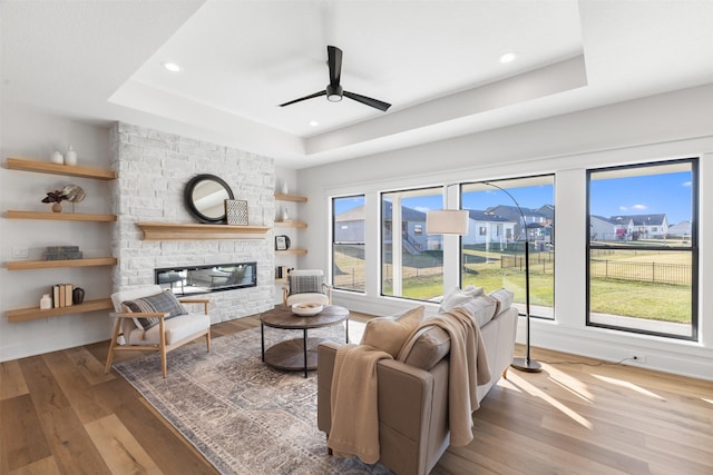 living room featuring hardwood / wood-style floors, ceiling fan, a stone fireplace, and a tray ceiling