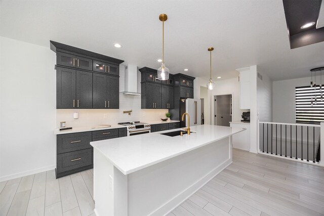 kitchen with an island with sink, hanging light fixtures, sink, wall chimney exhaust hood, and stainless steel appliances