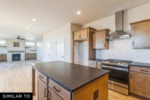 kitchen featuring ceiling fan, a center island, wall chimney exhaust hood, stainless steel gas range oven, and a stone fireplace