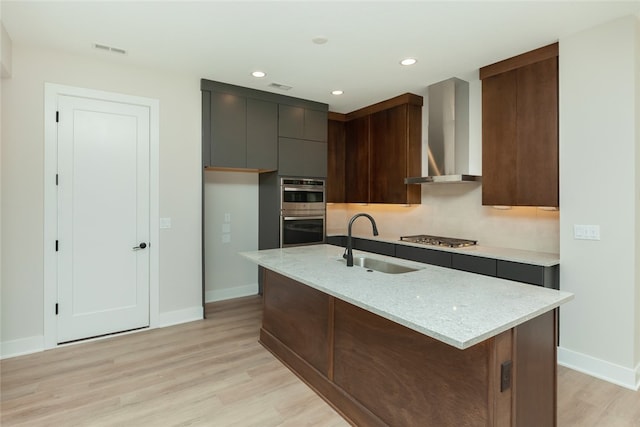 kitchen featuring light stone counters, sink, a center island with sink, wall chimney range hood, and light hardwood / wood-style floors