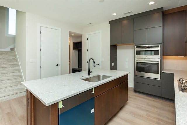 kitchen featuring sink, a kitchen island with sink, light hardwood / wood-style flooring, stainless steel double oven, and light stone countertops