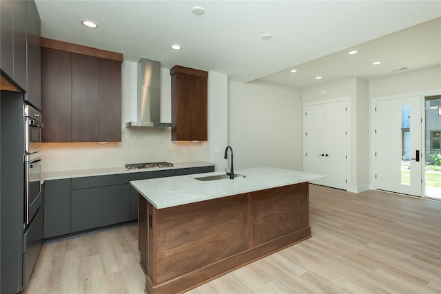 kitchen with wall chimney range hood, a kitchen island with sink, light hardwood / wood-style flooring, stainless steel gas stovetop, and sink