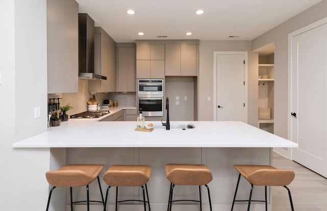 kitchen featuring kitchen peninsula, appliances with stainless steel finishes, wall chimney exhaust hood, gray cabinetry, and a kitchen bar