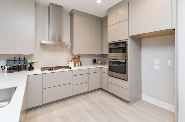 kitchen with appliances with stainless steel finishes, wall chimney exhaust hood, light wood-type flooring, tasteful backsplash, and gray cabinetry