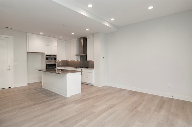 kitchen featuring light wood-type flooring, white cabinets, wall chimney exhaust hood, stainless steel double oven, and a center island with sink