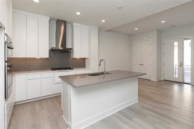 kitchen with wall chimney exhaust hood, a kitchen island with sink, light wood-type flooring, sink, and white cabinetry