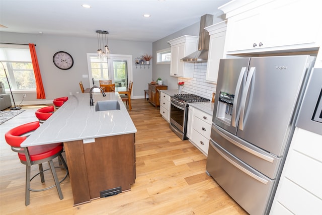 kitchen featuring sink, a center island with sink, wall chimney range hood, appliances with stainless steel finishes, and light hardwood / wood-style floors