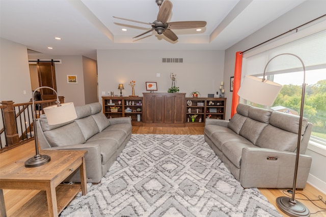 living room featuring a barn door, light hardwood / wood-style floors, a tray ceiling, and ceiling fan