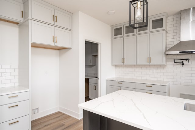 kitchen with light wood-type flooring, light stone countertops, wall chimney exhaust hood, hanging light fixtures, and backsplash