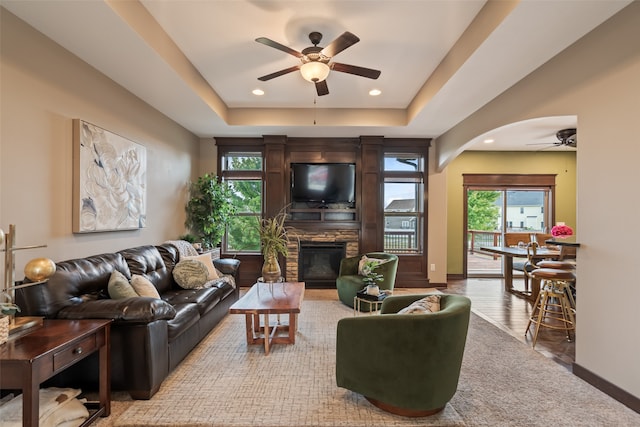 living room featuring a raised ceiling, wood-type flooring, ceiling fan, and a stone fireplace