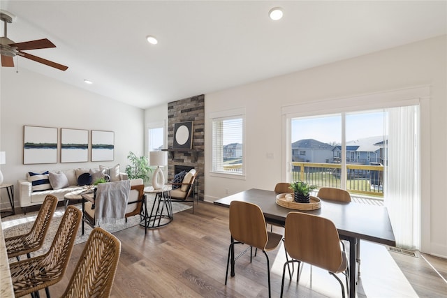 dining space with ceiling fan, a stone fireplace, light wood-type flooring, and vaulted ceiling