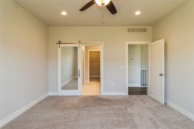 unfurnished bedroom featuring a spacious closet, light carpet, a barn door, and ceiling fan