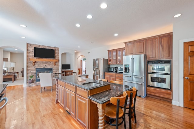 kitchen featuring a stone fireplace, an island with sink, dark stone counters, appliances with stainless steel finishes, and light hardwood / wood-style floors