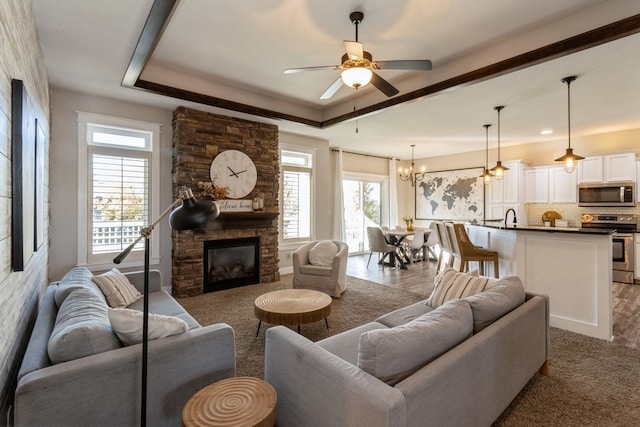 living room featuring sink, ceiling fan with notable chandelier, a fireplace, a tray ceiling, and dark wood-type flooring