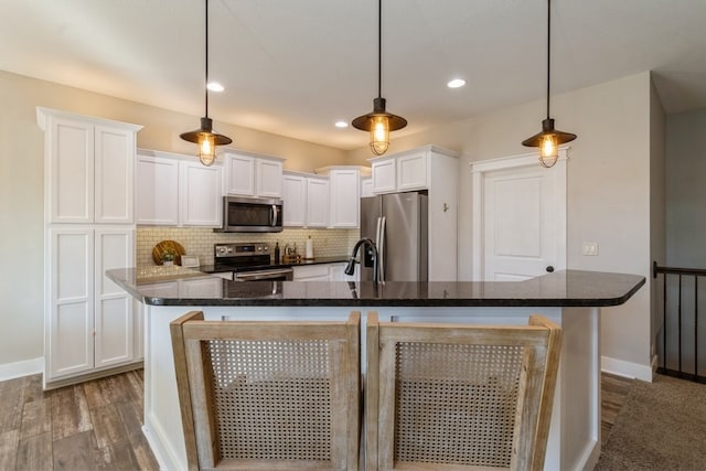 kitchen with a center island with sink, white cabinetry, wood-type flooring, stainless steel appliances, and decorative light fixtures