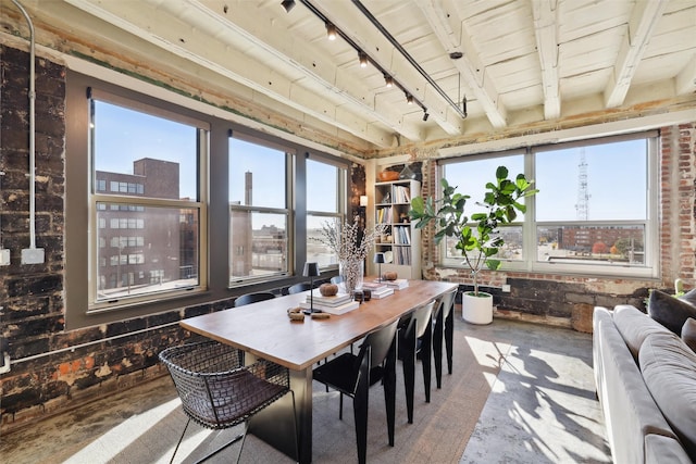 dining space featuring beamed ceiling, track lighting, plenty of natural light, and brick wall