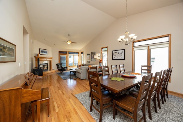 dining area featuring light wood-style flooring, high vaulted ceiling, a tile fireplace, baseboards, and ceiling fan with notable chandelier