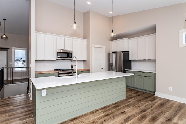 kitchen featuring white cabinets, decorative light fixtures, stainless steel appliances, a kitchen island with sink, and green cabinets