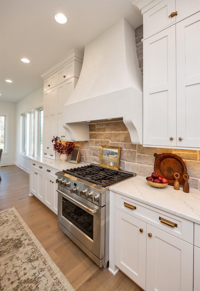 kitchen with white cabinets, high end stainless steel range, custom exhaust hood, light wood finished floors, and tasteful backsplash