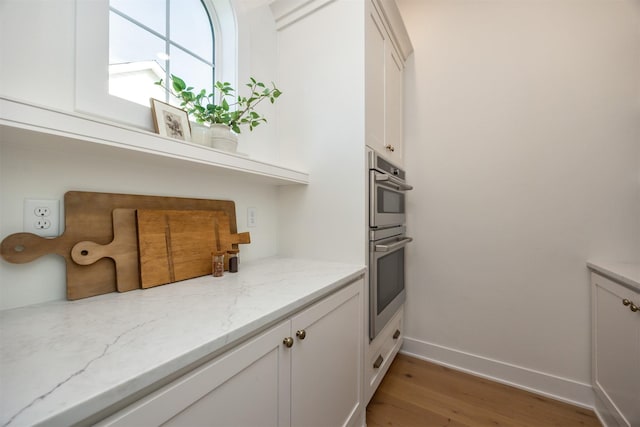 kitchen featuring light wood finished floors, baseboards, light stone counters, stainless steel double oven, and white cabinetry