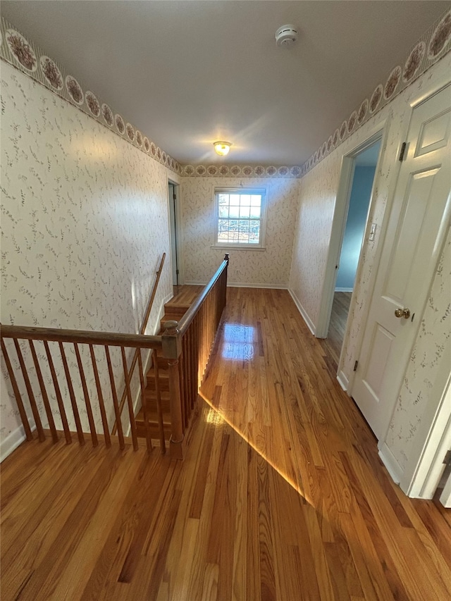 corridor with baseboards, wallpapered walls, wood finished floors, and an upstairs landing