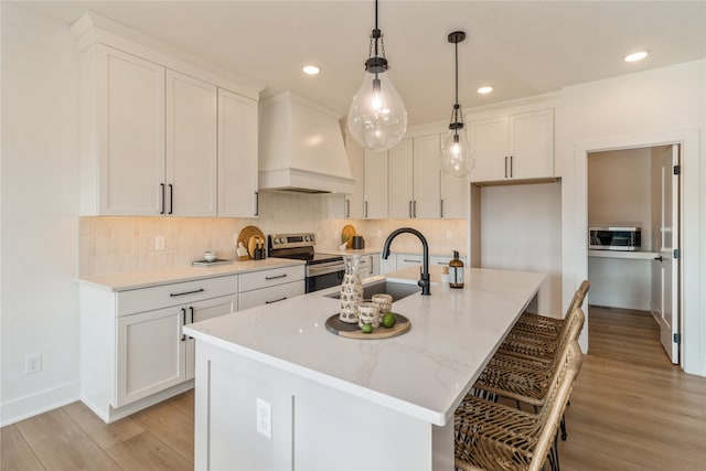 kitchen featuring an island with sink, sink, premium range hood, white cabinets, and stainless steel electric range oven