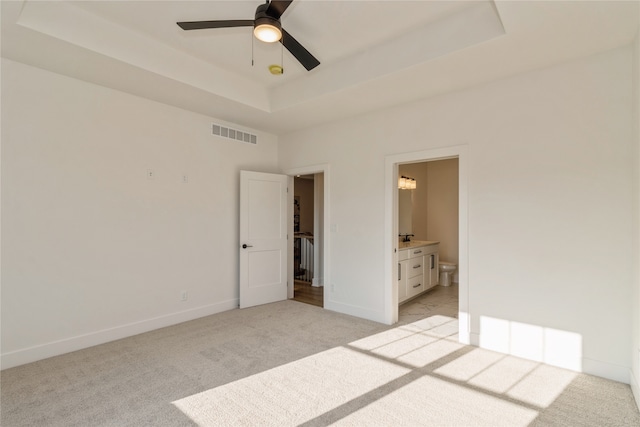 unfurnished bedroom featuring ensuite bath, light colored carpet, ceiling fan, and a raised ceiling