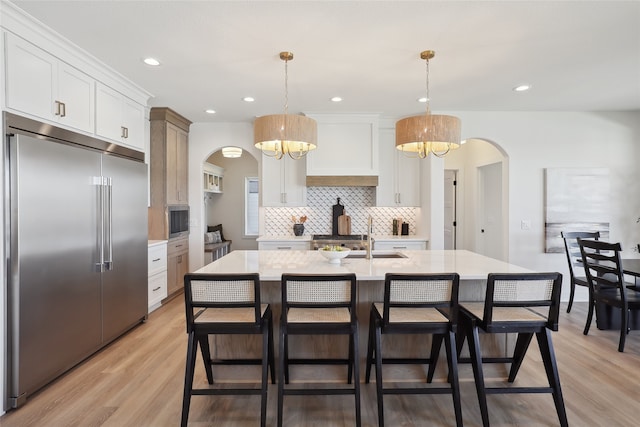 kitchen featuring built in appliances, light hardwood / wood-style flooring, a center island with sink, pendant lighting, and white cabinets