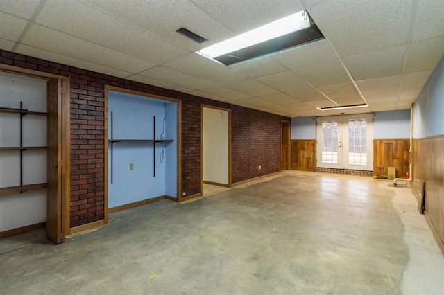 basement featuring french doors, a drop ceiling, wooden walls, and brick wall