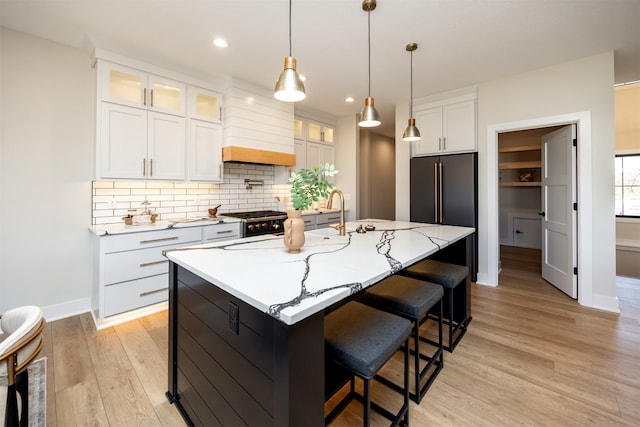 kitchen with custom exhaust hood, decorative backsplash, white cabinetry, a kitchen island with sink, and light wood-type flooring