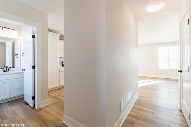 hall with visible vents, light wood-style flooring, a sink, a textured ceiling, and baseboards