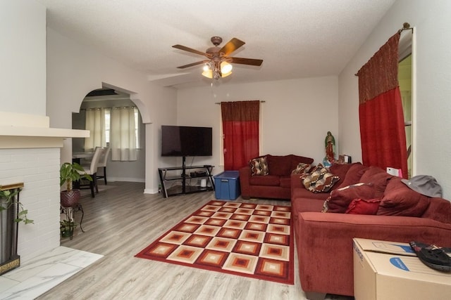 living room with a brick fireplace, ceiling fan, a textured ceiling, and light hardwood / wood-style flooring