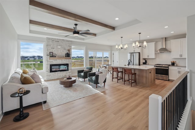 living room featuring light wood-type flooring, ceiling fan with notable chandelier, sink, beamed ceiling, and a fireplace