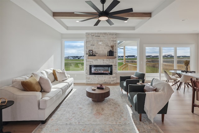 living room with beam ceiling, ceiling fan, a raised ceiling, a stone fireplace, and hardwood / wood-style floors