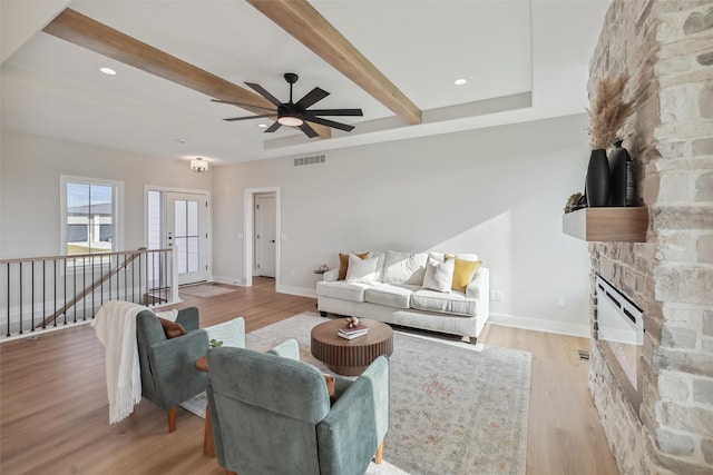 living room featuring beamed ceiling, light hardwood / wood-style flooring, ceiling fan, and a stone fireplace