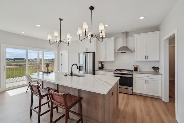 kitchen with wall chimney range hood, an inviting chandelier, a kitchen island with sink, white cabinets, and appliances with stainless steel finishes