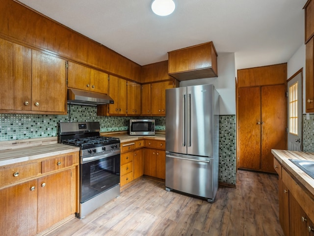 kitchen featuring under cabinet range hood, stainless steel appliances, brown cabinetry, and light countertops