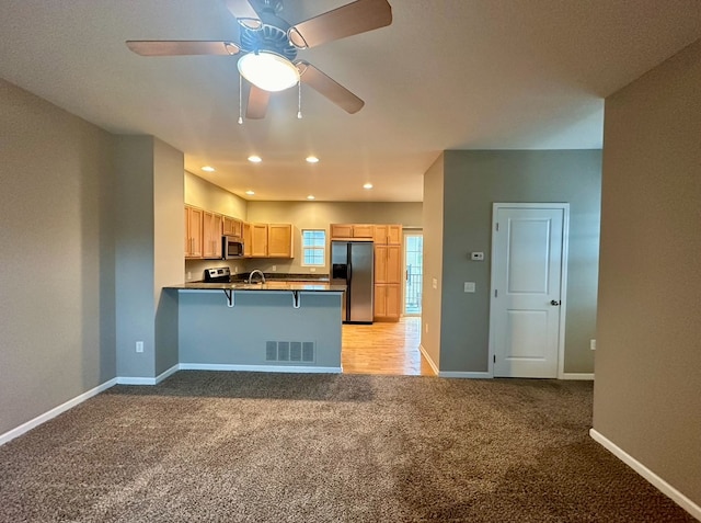 kitchen with light brown cabinets, kitchen peninsula, light colored carpet, appliances with stainless steel finishes, and ceiling fan