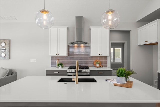 kitchen with stainless steel gas range oven, a center island with sink, wall chimney range hood, decorative light fixtures, and white cabinetry