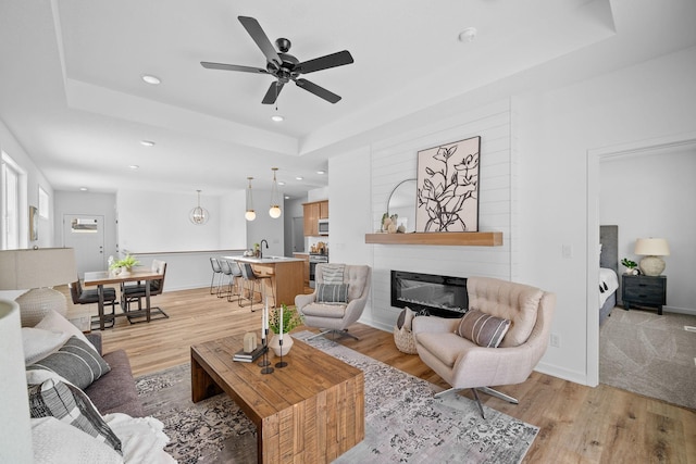 living room with sink, ceiling fan, a fireplace, a tray ceiling, and light hardwood / wood-style floors