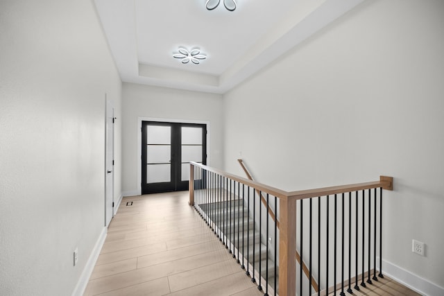 hall featuring baseboards, french doors, light wood-style flooring, a tray ceiling, and an upstairs landing