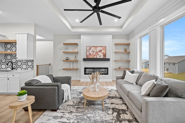 living area with ceiling fan, a raised ceiling, light wood finished floors, and a glass covered fireplace