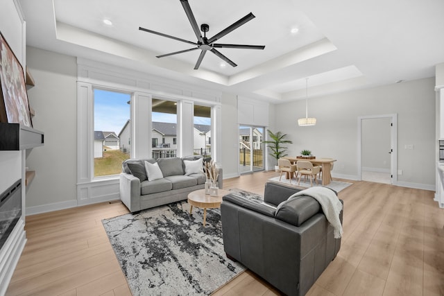 living room with a tray ceiling, light wood finished floors, a glass covered fireplace, ceiling fan, and baseboards